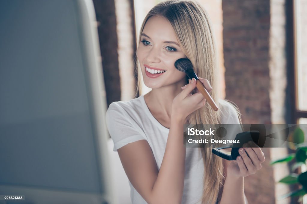 Attractive beautiful charming joyful glad woman wearing white t-shirt is applying blusher on her cheekbones in front of mirror at home Make-Up Stock Photo