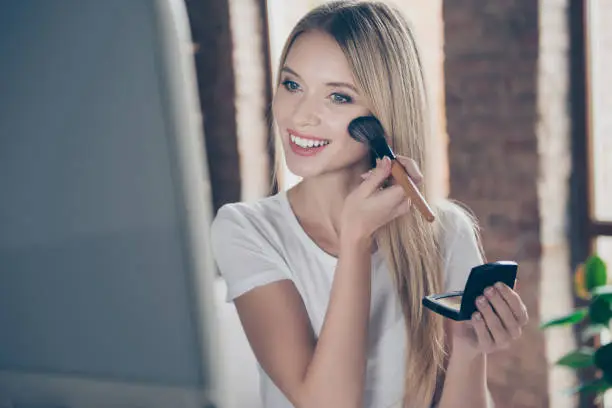 Photo of Attractive beautiful charming joyful glad woman wearing white t-shirt is applying blusher on her cheekbones in front of mirror at home