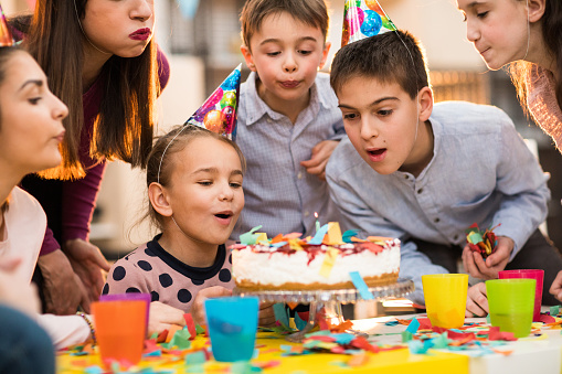 Group of children and an adult woman standing around the cake wearing party hats and feeling positive