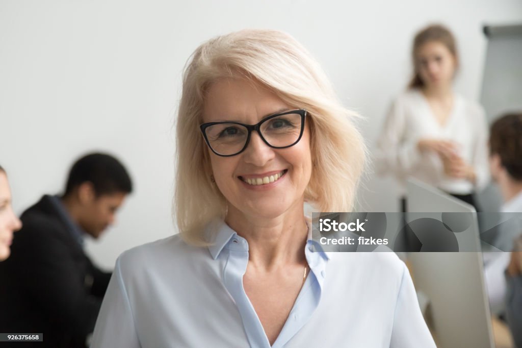 Smiling senior businesswoman wearing glasses portrait with businesspeople at background Portrait of smiling senior businesswoman wearing glasses with businesspeople at background, happy older team leader, female aged teacher professor or executive woman boss looking at camera, head shot Senior Adult Stock Photo