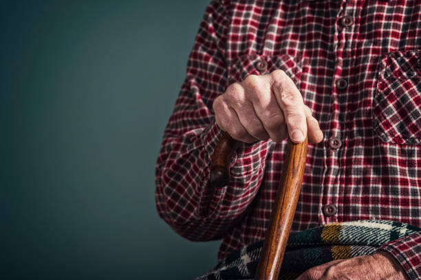 Close-up of an old Caucasian man's arthritic hand resting on top of an old wooden walking cane as he is sitting down inside his home. He wearing a red and black plaid flannel shirt.