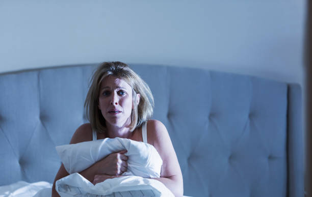 Mature woman waking up from bad dream A mature woman in her 40s sitting up in bed clutching her comforter with a worried look on her face. She looks like she had a bad dream. Or perhaps she is sick with the flu. horror waking up bed women stock pictures, royalty-free photos & images