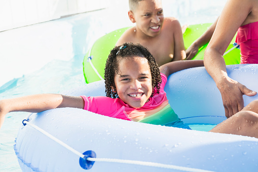A 7 year old girl with her family having fun at a water park on the lazy river.  She is mixed race African-American and Hispanic. She is in an inflatable innertube smiling at the camera.