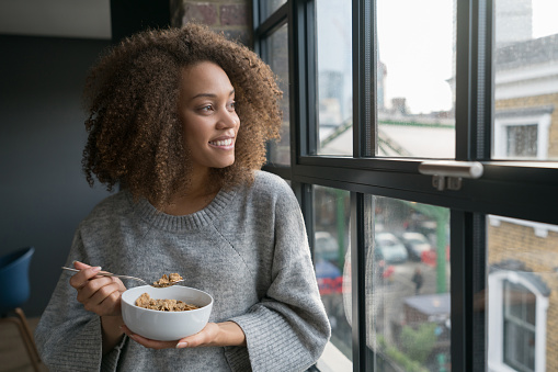Portrait of a black woman eating a bowl of cereals at home and looking through the window smiling - lifestyle concepts