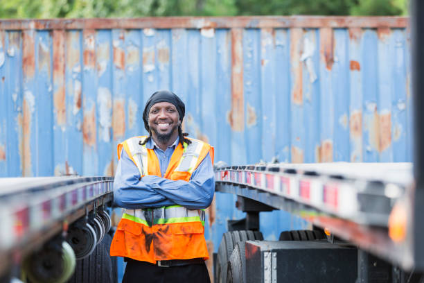 Mature black man in safety vest next to his truck A mature black man in his 40s wearing a reflective vest, looking at the camera, arms crossed. He is a truck driver or worker in a distribution warehouse, standing between two semi-trucks. do rag stock pictures, royalty-free photos & images