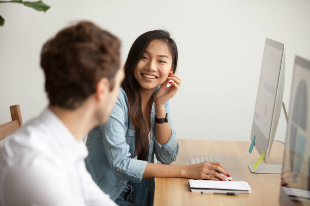 Smiling attractive asian woman talking to male colleague at work Smiling attractive asian woman talking to male colleague at work sharing office desk with desktops, friendly multiracial coworkers interns having pleasant fun conversation at workplace together work romance stock pictures, royalty-free photos & images