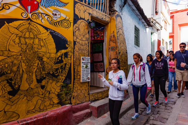 Bogotá, Colombia - Local Colombians And Tourists On The Cobblestoned Calle del Embudo, In The Historic La Candelaria District of The Andean Capital City Bogota, Colombia - May 28, 2017: Local Colombian people walk down the narrow Calle del Embudo which gets its name from its shape. The English translation of the name would be, "Funnel Street." At this end it is at its narrowest. It is obvious that only two people can At the same time can walk down the street at this point. It is one of the most colorful streets in the historic La Candelaria district of Bogotá, the Andean capital city of the South American country of Colombia. Constructed over 450 years ago, the street leads to the Chorro de Quevedo, the plaza where it is believed the Spanish Conquistador, Gonzalo Jiménez de Quesada founded the city in 1538. Many street facing walls in this area are painted with either street art or the legends of the Pre-Colombian era, in the vibrant colours of Colombia. Photo shot in the afternoon sunlight; horizontal format. Camera: Canon EOS 5D MII. Lens EF 24-70 mm F2.8L USM. calle del embudo stock pictures, royalty-free photos & images
