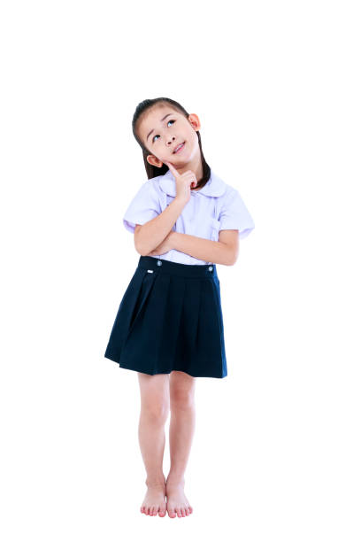 Full body of preschool girl in uniform smiling and looking up, her index finger at cheek. Cute asian thoughtful child standing at studio. Isolated on white background. Education and people concept.