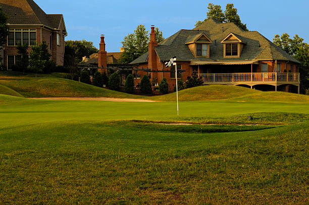 this color image is of a Putting Green on Professional Golf Course near Luxury Homes. the putting green has manicured grass or lawn with two sand traps around the outside of the putting green and putting hole. the grass is green and their are expensive houses or homes in the background. the photo was taken during the spring or summer. and the lighting is natural sunlight during the evening part of the day. there are also trees with leaves in the background. 