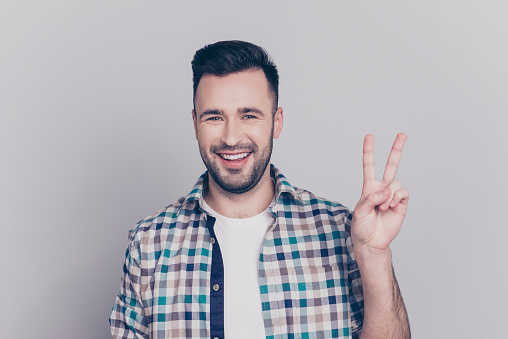 Portrait of young, smiling man in checkered shirt with stubble, bristle gesturing v-sign over grey background