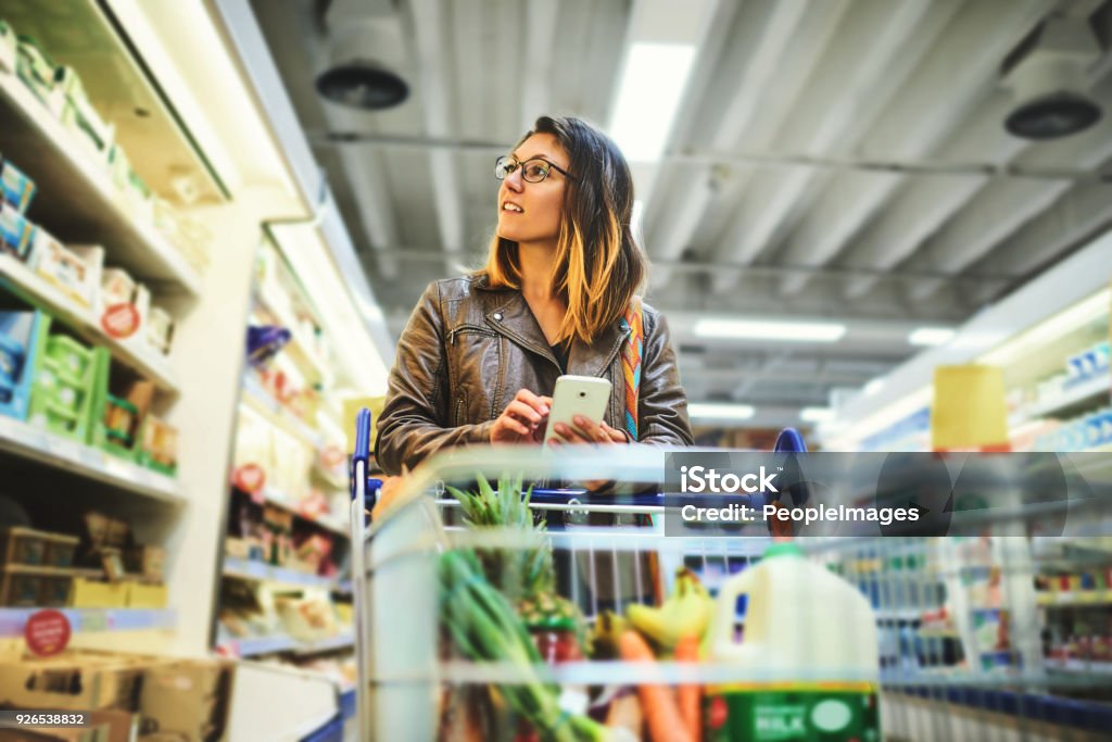 Shopping lists in app format Shot of a young woman using a mobile phone in a grocery store Supermarket Stock Photo