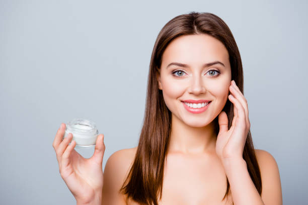 istock Concept of using moisturizing cream before going to bed. Beautiful cute pretty charming woman is holding a cream jar and applying it on her skin, isolated on grey background 926546288