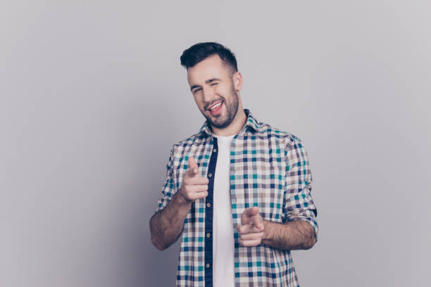 Isolated confident, smiling man in checkered shirt pointing towards his forefingers and winking with one eye to camera over grey background Isolated confident, smiling man in checkered shirt pointing towards his forefingers and winking with one eye to camera over grey background young man wink stock pictures, royalty-free photos & images