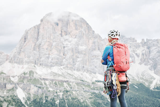 Female mountaineer with backpack, helmet and harness with climbing gear enjoying stunning view to mount Tofana di Rozes before ascent during summer day in Dolomite Alps - adventure concept