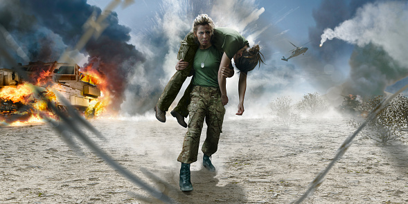 A female army medic dressed in camouflage fatigues carrying a wounded female soldier on her shoulders in a firemans lift away from danger on an active desert battlefield. A generic army truck is burning in the background, a helicopter in flight and the sky full of smoke and explosions.