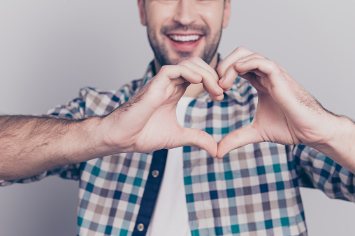 Close up portrait of heart made from fingers of man in checkered shirt who prepared, sending love symbol to his girlfriend over grey background