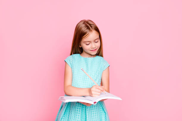 bella adorabile bambina affascinante sta guardando il libro di copie tra le sue mani e scrivendo informazioni lì, indossa un vestito azzurro, isolato su sfondo rosa brillante, copyspace - schoolgirl school children isolated child foto e immagini stock