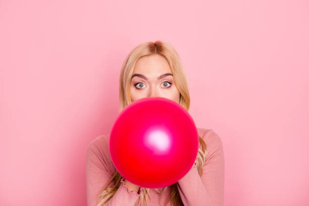 bouchent portrait d’une femme drôle en gonflant un ballon rouge pour partie avec les yeux grands ouverts et sourcils surélevés, debout sur fond rose - blowing a balloon photos et images de collection