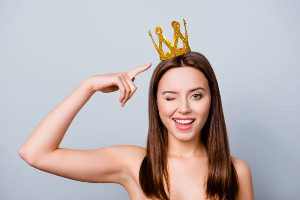 istock Confident cute beautiful young woman with a crown on her head is thinking that she is a queen, she is pointing on her crown, isolated on grey background 926558334