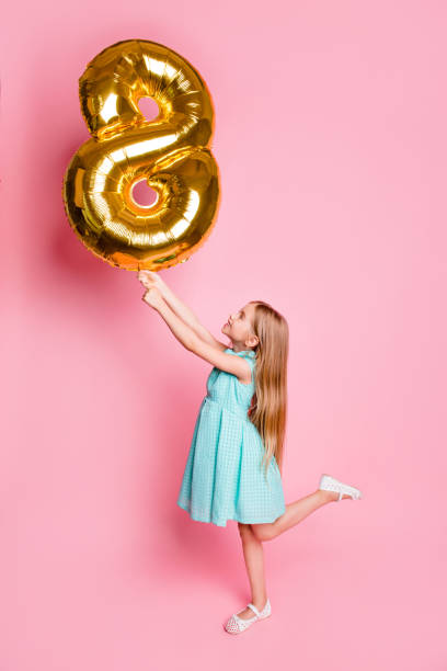 i am 8 years old! vertical full-length side profile view portrait of cute lovely beautiful girl celebrating birthday, she is holding golden balloon in shape of figure-eight isolated on pink background - personal accessory balloon beauty birthday imagens e fotografias de stock