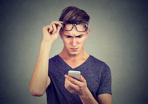Casual confused young man in t-shirt and eyeglasses surfing smartphone on gray background