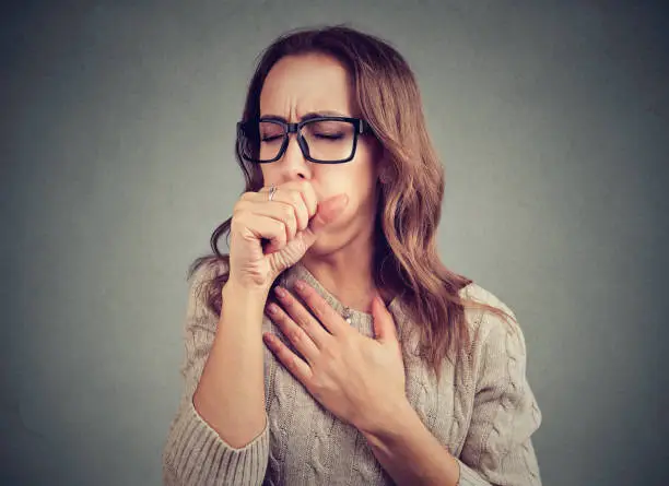 Young woman having asthma problems and coughing badly covering mouth and holding hand on chest.