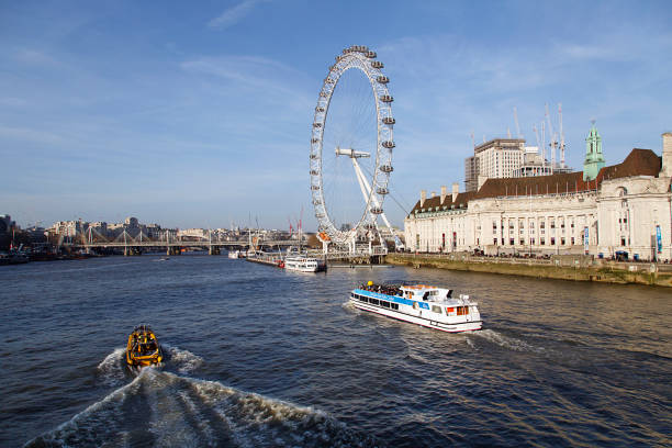 London Eye River Cruise boat passes the Millennium Wheel London, UK: February 24, 2018: London Eye River Cruise boat passes the Millennium Wheel on the Thames River. Cruising the Thames is a leisurely way to view the new and historic buildings along the embankment on the way to Greenwich. london county hall stock pictures, royalty-free photos & images
