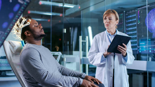 istock Man Wearing Brainwave Scanning Headset Sits in a Chair while Scientist with Tablet Computer Supervises Process. In the Modern Brain Study Laboratory Monitors Show EEG Reading and Brain Model. 926573756