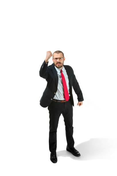 Angry aggressive businessman threatening by fist to camera. Isolated on white studio background. Serious bearded young man in glasses, suit, red tie. Business, threat, boss concept.