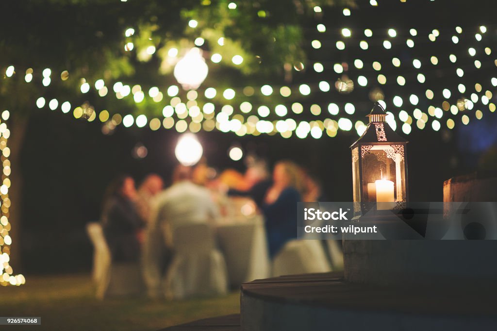 Candle and string lights outdoor dinner A string of fairy lights and a chandelier with candle hanging in-between trees photographed shining at night with image focus technique focus on the foreground bokeh background and dark and a table with peopleFranschhoek Cape Winelands South Africa Outdoors Stock Photo