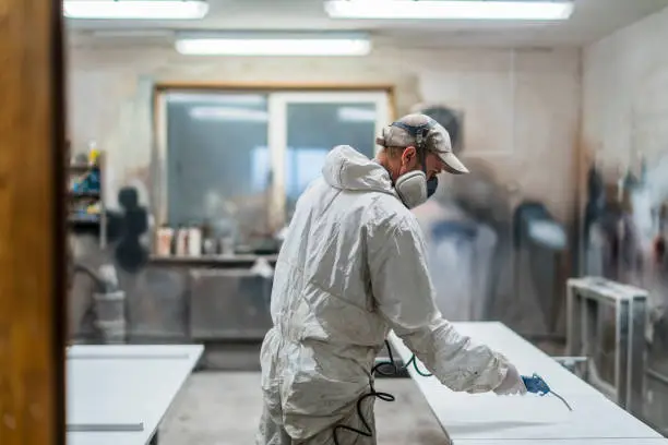 The mature man, manual worker, wearing the protective mask and coveralls, cleaning surfaces for painting with the compressed air gun at the small furniture manufacture