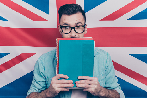 English language learning concept-portrait of excited man holding colorful copy books in hands closing half face with notebooks standing over English flag background