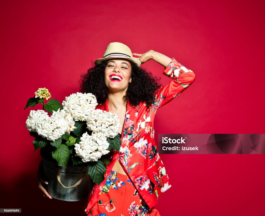 Summer portrait of excited young woman with flowers against red background Summer portrait of beautiful, excited young woman wearing red floral pattern jacket and sunhat, standing against red background, holding flowers. Gardening Stock Photo