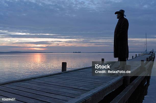 Silhouette Of Man Standing On Lakeside Jetty At Sunset Stock Photo - Download Image Now