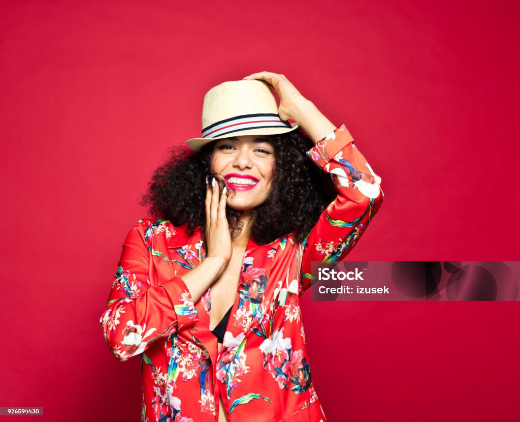 Summer portrait of excited young woman against red background Summer portrait of beautiful, excited young woman wearing red floral pattern jacket and sunhat, standing against red background. Laughing Stock Photo