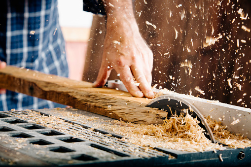 Male carpenter using saw in home workshop with wood chips flying