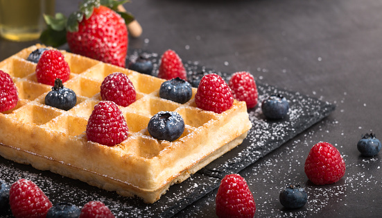 Freshly baked Brussels waffles on a black stone plate with fresh strawberry and blueberry and raspberry on a dark stone table with honey and coffee.