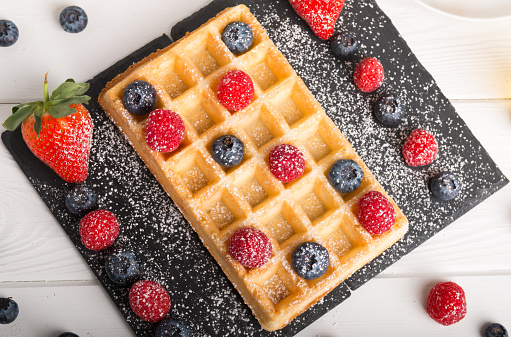 Freshly baked Brussels waffles on a black stone plate with fresh strawberry and blueberry and raspberry on a white wooden table with honey and coffee.