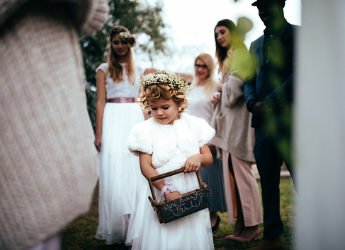 Flower girl holding basket and walking down the aisle with bride at outdoors wedding ceremony