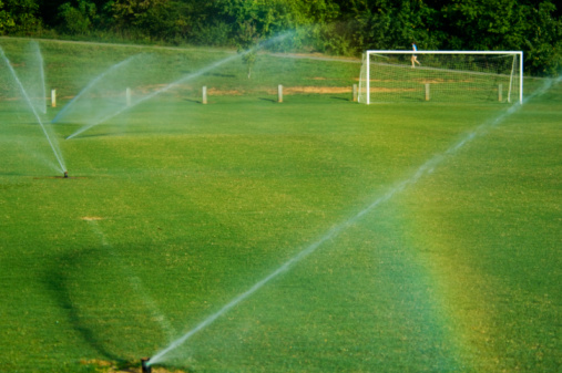 this photo is of a Soccer Field with Soccer Goal at Park. the soccer field is lush green grass, mowed lawn or artificial turf. the soccer goal is white with a soccer net. the water sprinklers are watering the grass in the morning. and there is a rainbow in the picture. the soccer field is at a local park or school. and there are trees in the background. and the picture was taken during the day during the spring or summer. and the lighting is natural sunlight. 