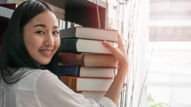 istock Young Asian female university student hold stack of textbooks with both hands in front of bookshelf in university library 926637608