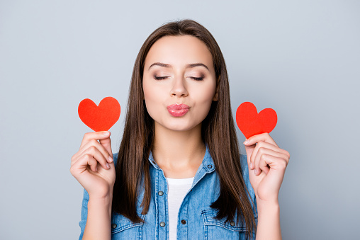 Close up portrait of brunette girl, holding two small red hearts in her raised hands, blowing kiss to the camera with closed eyes, dreaming about her lover while standing over grey background
