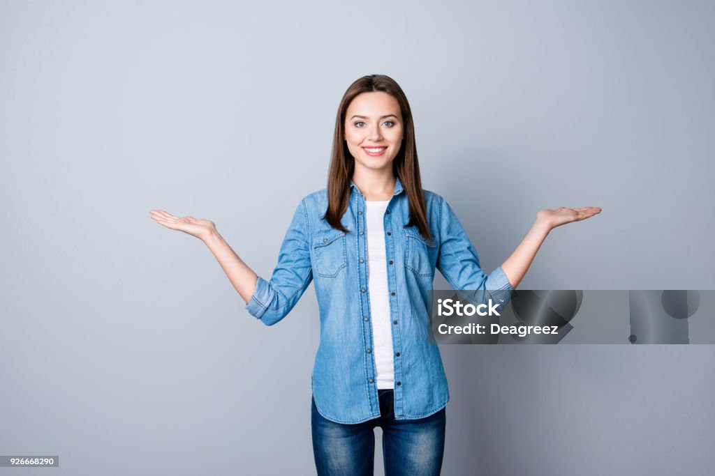 Portrait of successful cheerful cute young woman presenting something, showing copy space on her palm in two sides, looking at camera, standing over grey background Two Objects Stock Photo