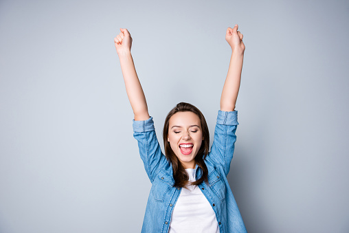 Amazed brunette young business woman in casual shirt is gesturing victory with her raised hands, she is shocked, extremely happy, with closed eyes, beaming smile, open mouth  on  grey background
