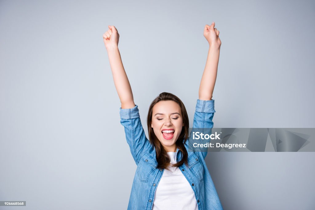 Amazed brunette young business woman in casual shirt is gesturing victory with her raised hands, she is shocked, extremely happy, with closed eyes, beaming smile, open mouth  on  grey background Women Stock Photo