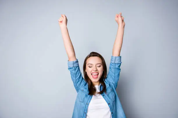 Photo of Amazed brunette young business woman in casual shirt is gesturing victory with her raised hands, she is shocked, extremely happy, with closed eyes, beaming smile, open mouth  on  grey background