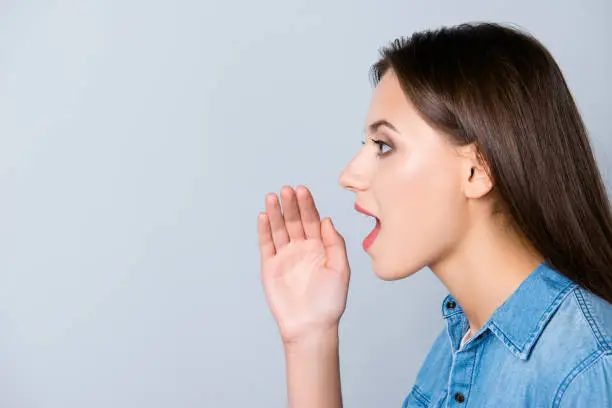 Hey, listen here! Close up side-view portrait of attractive woman telling news, shouting and holding hand near her open mouth, standing over grey background with copy space