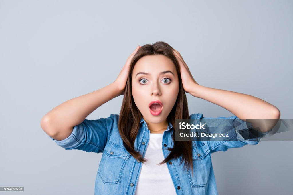 Omg! Close up portrait of amazed girl with wide open mouth and eyes in casual wear, holding head with her hands, wondering what happened, looking confused, lost on the grey background One Woman Only Stock Photo