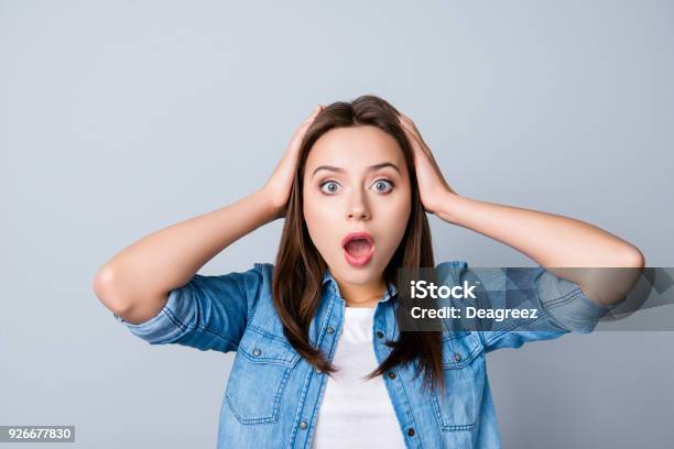Omg Close Up Portrait Of Amazed Girl With Wide Open Mouth And Eyes In Casual Wear Holding Head With Her Hands Wondering What Happened Looking Confused Lost On The Grey Background Stock Photo - Download Image Now