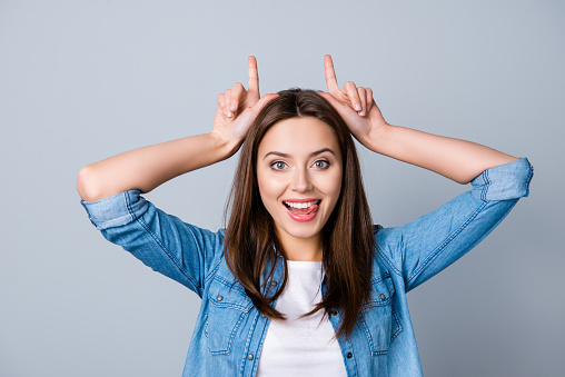 Close up portrait of a nice, smiling girl  with raised hands holding two fingers as antlers on head, looking very funny, showing her tongue, standing over grey background